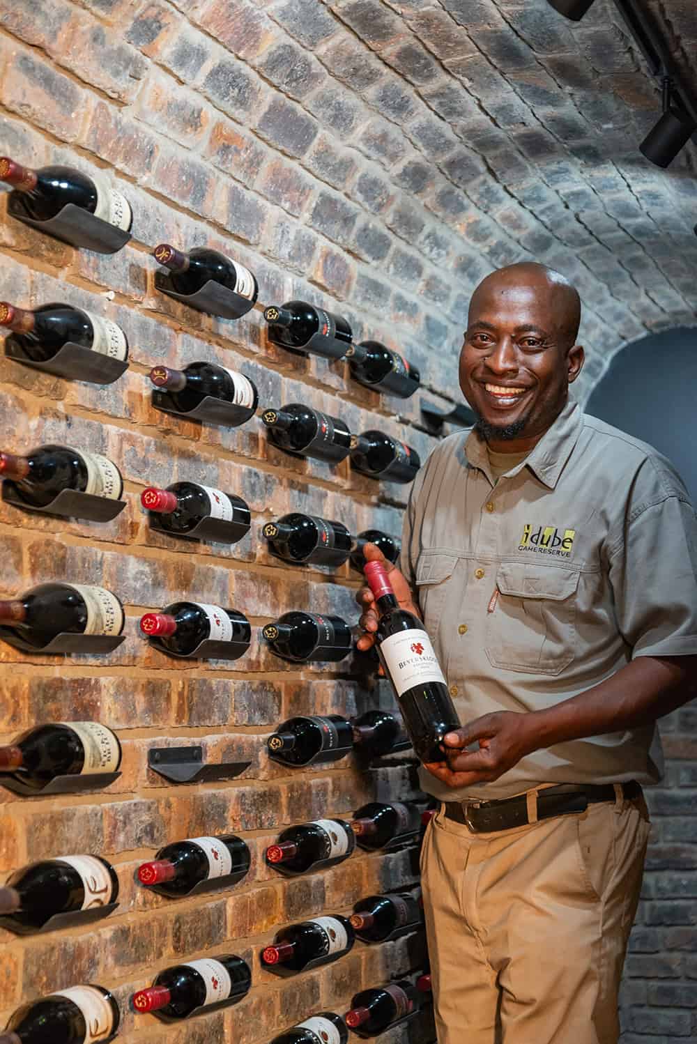 Wine tasting at Idube Game Reserve, with a staff member selecting a bottle against a brick wall backdrop.